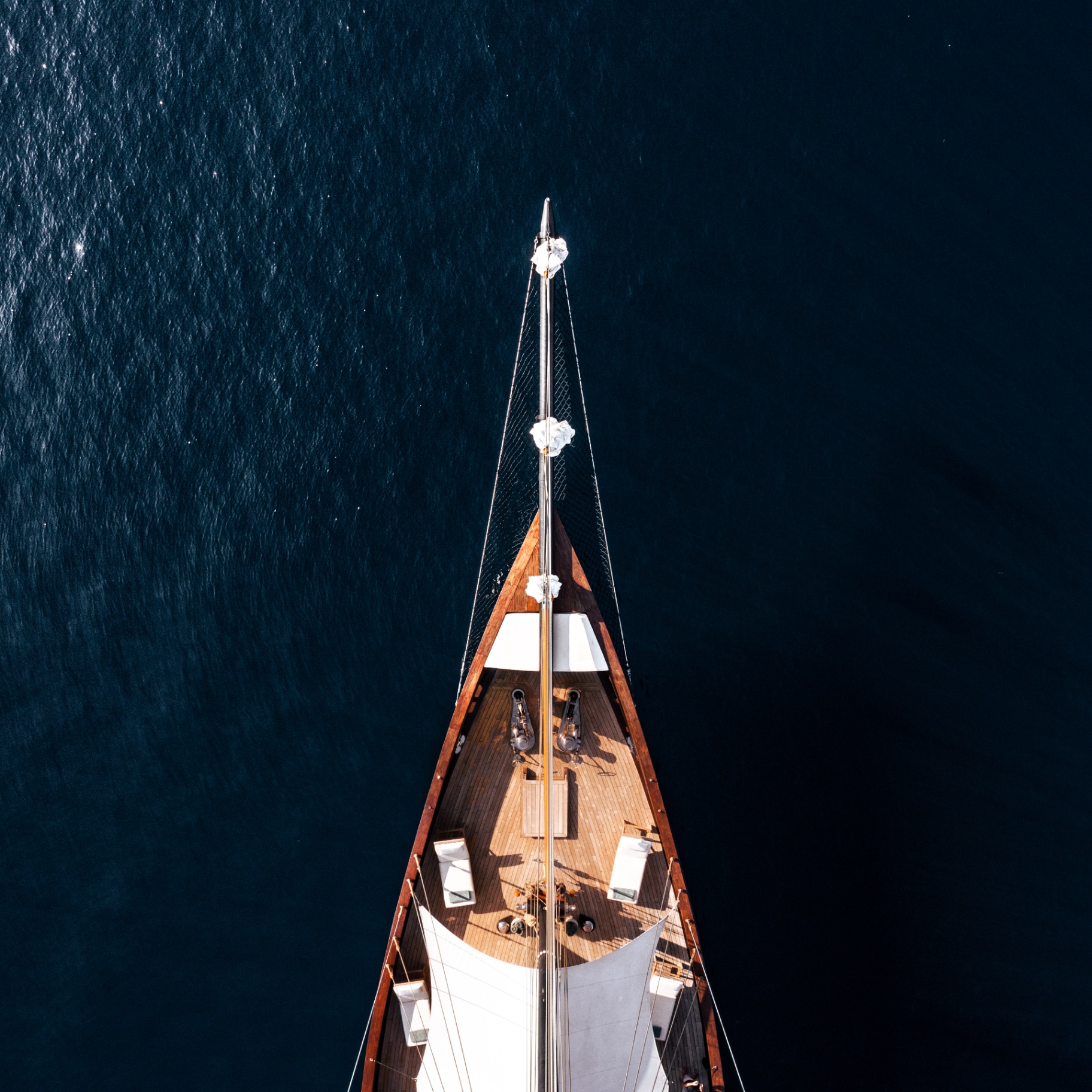 Aerial top-down of Vela's bow, mast and teak foredeck over deep blue water