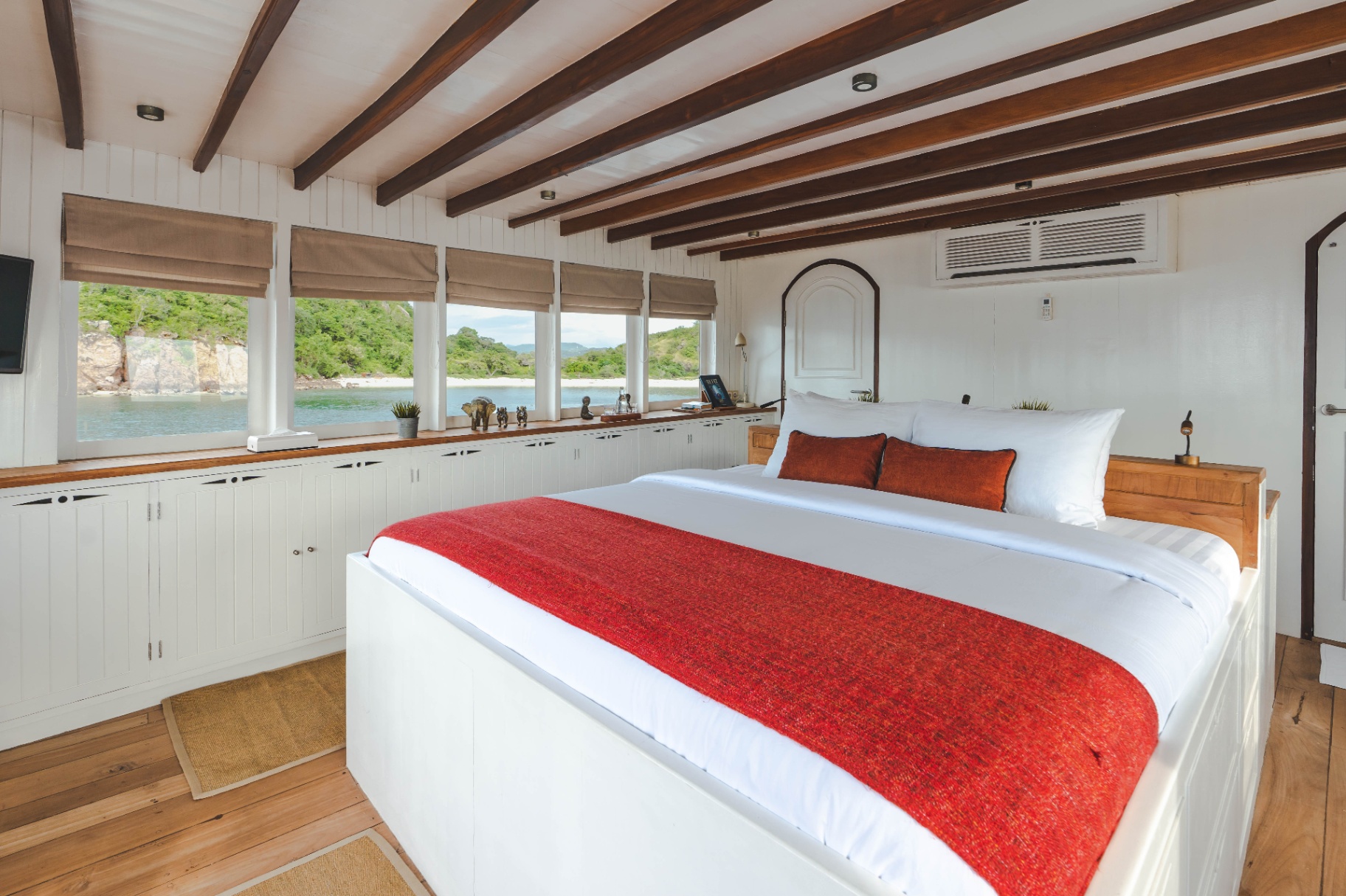 Komodo suite bed and reading nook on Mischief with red throw, wall-mounted TV and full-length windows over the sea