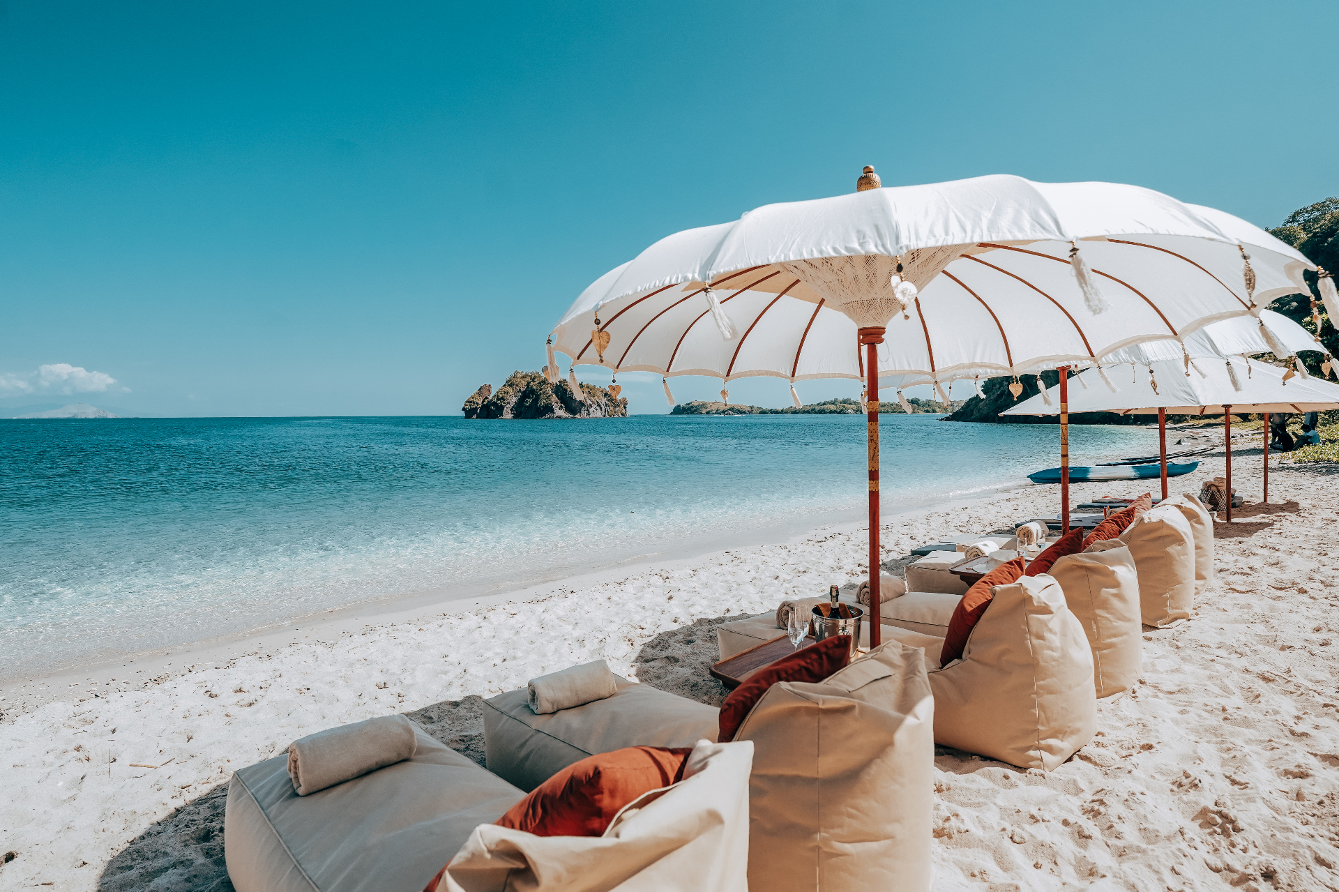 Row of cream beanbag loungers with orange cushions and a large white parasol on a white-sand beach beside turquoise water