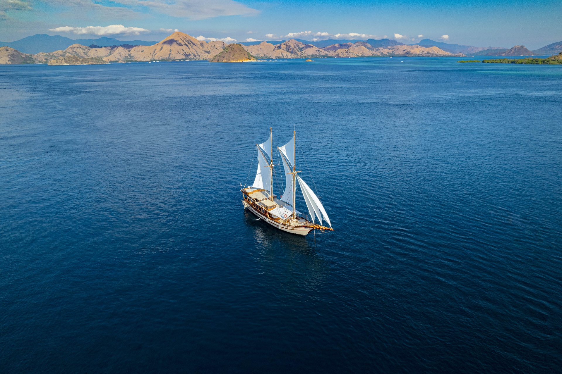 Aerial of Nataraja sailing across deep blue water with Komodo's dry hills in the distance