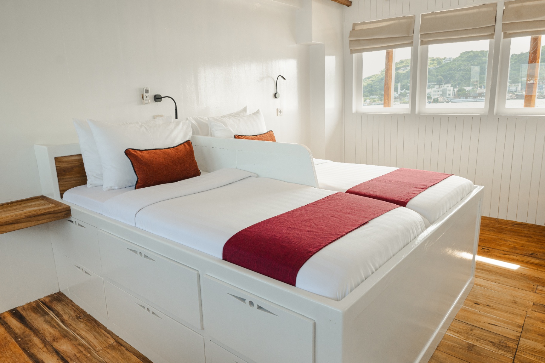 Mischief Banda cabin with king bed, orange accent cushions, teak floor and porthole-style windows over the harbour