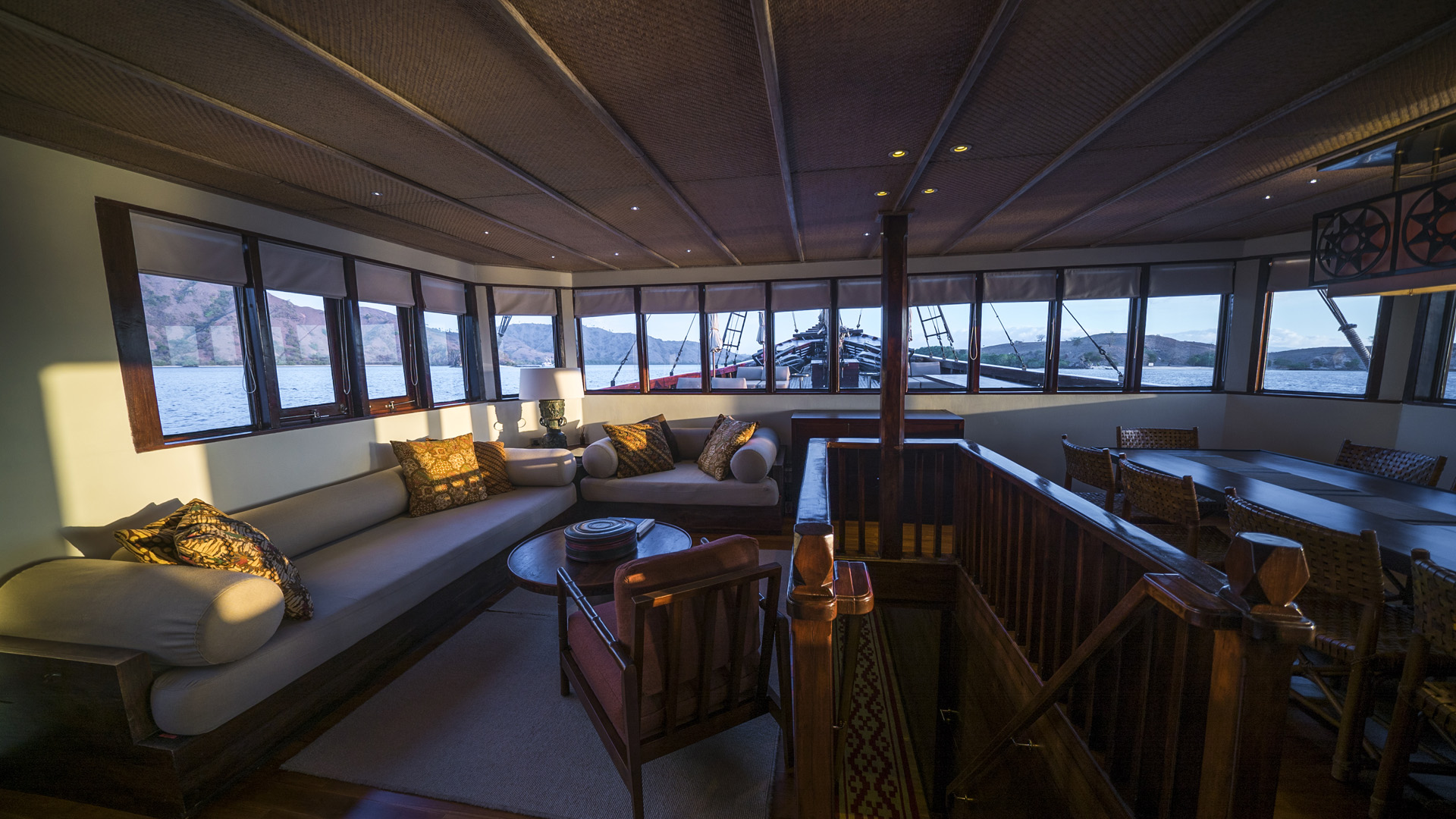 Main-deck lounge on Alila Purnama at golden hour with panoramic windows over Komodo