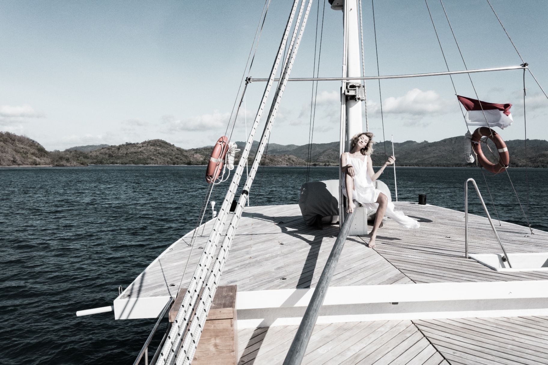 Woman in a white dress seated on Splendour's foredeck with the Indonesian flag and green hills behind