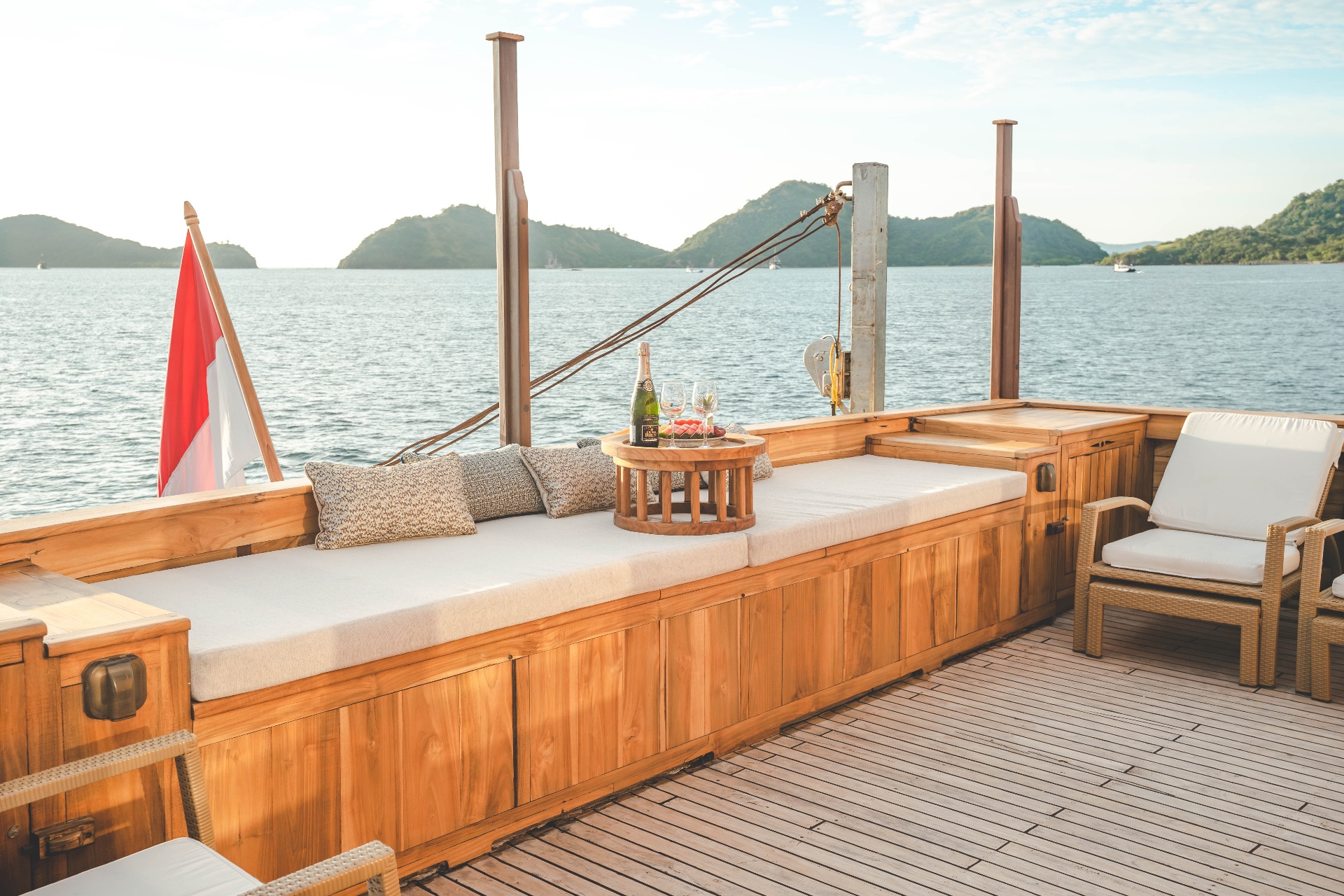 Mischief bow daybed with pillows, champagne and fruit tray, framed by the Indonesian flag and Komodo hills